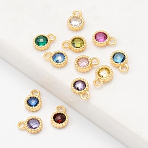 Könnte beinhalten: Eine Sammlung kleiner, goldfarbener Charms mit farbigen, facettierten Edelsteinen. Die Charms verfügen über einen runden Edelstein in verschiedenen Farben, darunter Grün, Blau, Rosa und Lila, der in einer goldfarbenen Lünette mit einer kleinen Schlaufe gefasst ist.