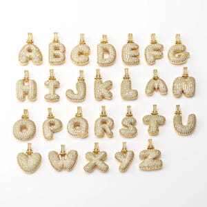 May include: A set of 26 gold-plated alphabet letter pendants, each letter is encrusted with clear cubic zirconia stones. The letters are in a bubble font style.