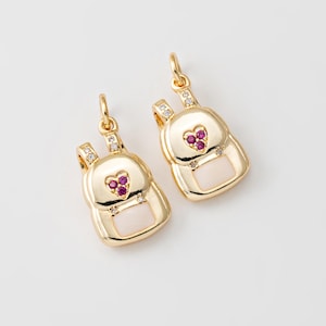 May include: Two gold-tone backpack charms with a white enamel background and pink heart accents. Each charm features a small heart with three pink gemstones.