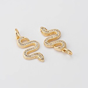 Op de afbeelding: Twee gouden slangencharms met heldere kristallen accenten. De charms zijn in de vorm van een slang met zijn lichaam gekruld in een cirkel.