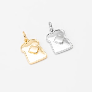 Könnte beinhalten: Zwei Anhänger, einer goldfarben und einer silberfarben, jeweils in Form einer Toastscheibe mit einem Butterquadrat. Die Anhänger befinden sich auf weißem Hintergrund. Der goldene Anhänger hat eine Schlaufe für eine Kette.
