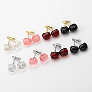 Op de afbeelding: Assortiment van kersen-vormige bedels in verschillende kleuren, waaronder helder, roze, rood en zwart. De bedels zijn gemaakt van glas en hebben een gouden, zilveren of zwarte metalen steel.