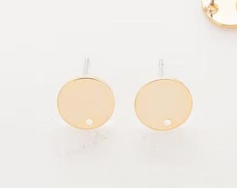 May include: A collection of gold-colored, round earring posts with a flat surface and a post for attaching to the ear. The earrings are scattered on a white surface, with two earrings isolated in the foreground.