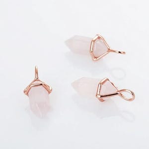 May include: Three rose quartz crystal pendants with gold-coloured metal accents. The crystals are light pink and have a pointed, hexagonal shape. The metal setting forms a loop for hanging.