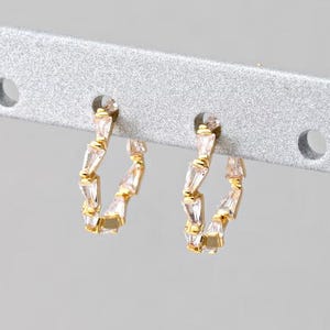 May include: A pair of gold-colored hoop earrings with clear, rectangular-cut stones. The earrings are displayed on a silver bar with two holes. The stones are set in a zig-zag pattern around the hoops, creating a sparkling effect.