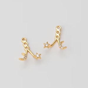 Puede incluir: Un par de pendientes dorados con un diseño celestial. Cada pendiente tiene una luna creciente y una estrella, ambas adornadas con pequeñas gemas transparentes. Los pendientes tienen cierre de poste y están sobre un fondo gris claro.