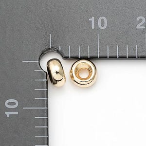 Puede incluir: Dos cuentas redondas doradas con un agujero en el centro. Las cuentas están junto a una regla con marcas en centímetros. Las cuentas son probablemente para la fabricación de joyas o manualidades.