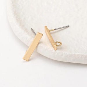 May include: Two gold-colored bar earrings with a post and loop design. The earrings are rectangular and have a matte finish. The posts are silver-colored. The earrings are displayed on a white, textured surface.