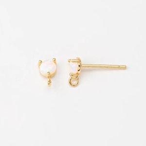 May include: A pair of gold-colored stud earrings with a small, round, white opal stone. The earrings have a post and a small loop for attaching charms or other decorative elements. The earrings are set against a white background.