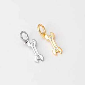 Könnte beinhalten: Zwei kleine Schraubenschlüssel-Anhänger, einer silberfarben und einer goldfarben, jeweils mit einem kleinen, klaren Edelstein. Die Anhänger befinden sich auf weißem Hintergrund. Der goldene Anhänger hat eine goldene Schlaufe, und der silberne Anhänger hat eine silberne Schlaufe.