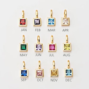 Op de afbeelding: Twaalf gouden bedels met verschillende vierkante edelstenen in verschillende kleuren, waarbij elke bedel een maand van het jaar voorstelt. De bedels zijn gelabeld met de drieliterige afkortingen voor elke maand: JAN, FEB, MAR, APR, MEI, JUN, JUL, AUG, SEP, OKT, NOV, DEC.