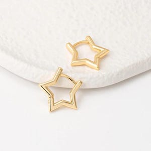 May include: Two gold-colored star-shaped hoop earrings. The earrings are made of a shiny metal and have a simple, modern design. The earrings are displayed on a white, textured surface.