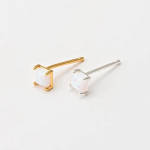May include: Two square-shaped stud earrings. One is gold-toned, the other is silver-toned. Each earring features a white, square-cut gemstone. The earrings are set against a white background.