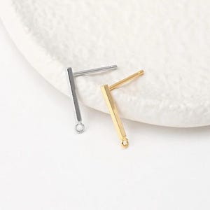 May include: Two minimalist bar stud earrings. One is silver, the other is gold. Each earring has a post for piercing and a small loop at the bottom. The earrings are displayed on a white, textured surface.
