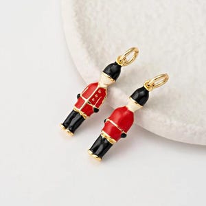 May include: Two gold-toned charms shaped like British Royal Guard figures. Each charm features a red jacket, black pants, and a black hat. The figures have white faces and gold accents. The charms are designed with a loop for attaching to jewelry.