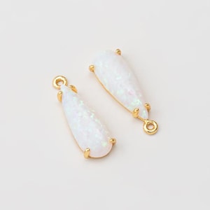 May include: Two teardrop-shaped pendants featuring iridescent white opal-like stones set in polished gold-tone metal frames. Each pendant has a small circular loop at the top and bottom, with four prongs securing the stone within the metal setting.