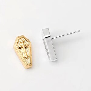 May include: A pair of coffin-shaped stud earrings. One earring is gold-colored with a cross design, and the other is silver-colored. The earrings are set against a white background.