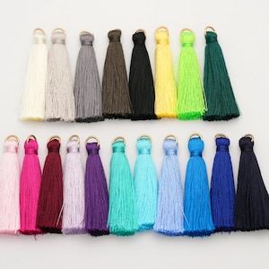 Puede incluir: Una colección de borlas coloridas en varios tonos, incluyendo rosa, rojo, morado, turquesa, azul, verde, amarillo, negro y gris. Cada borla tiene un lazo metálico dorado en la parte superior. Estas borlas decorativas son ideales para la creación de joyas o proyectos de artesanía.