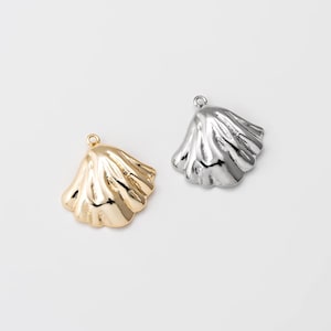 May include: Two seashell-shaped pendants, one gold-toned and one silver-toned, are displayed against a white background. Each pendant has a loop at the top for attaching to a chain or other jewelry component. The pendants have a textured, rippled surface.
