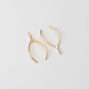 May include: Two gold-colored wishbone charms. The charms have a curved, wishbone shape with a decorative textured design at the top and a small loop for attaching to jewelry. The background is plain white.