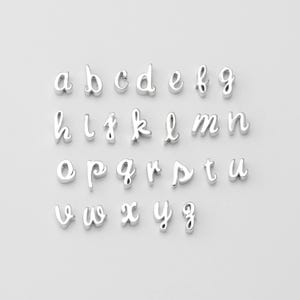 May include: A collection of silver-colored lowercase alphabet charms, each with a cursive font. The letters are arranged in rows, showcasing the entire English alphabet from a to z. Ideal for jewelry making or personalized crafts.