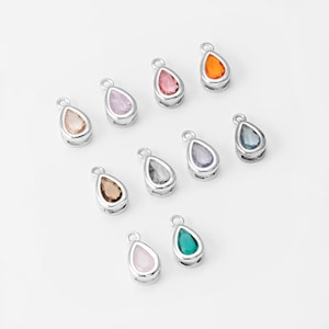 Könnte beinhalten: Eine Sammlung tropfenförmiger Anhänger mit silberfarbenen Metalleinfassungen. Jeder Anhänger hat einen andersfarbigen Edelstein, darunter rosa, orange, grün und blau. Die Anhänger sind auf einer weißen Oberfläche angeordnet.