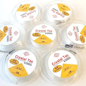 Puede incluir: Carretes de hilo elástico transparente para abalorios. Cada carrete tiene una etiqueta amarilla con el texto "Crystal Tec KOREA", medidas de diámetro y el texto "NEW STRONG". Los carretes miden 5 metros.