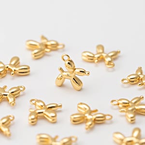 May include: Gold-colored balloon dog charms scattered on a white surface. Each charm is a miniature sculpture of a dog, with a loop for attaching to jewelry or other crafts. The charms have a shiny, metallic finish.