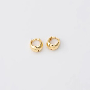 May include: A pair of small, gold-coloured hoop earrings. Each earring features a rounded, smooth design with a hinged closure. The earrings are presented on a white background, highlighting their polished, reflective surface.