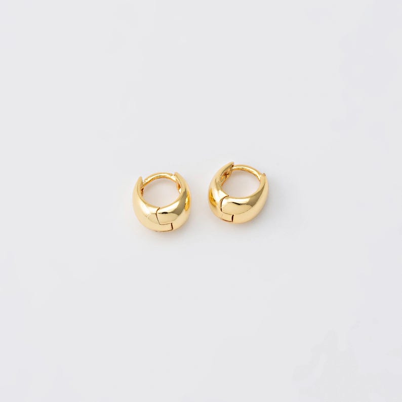 May include: A pair of small, gold-coloured hoop earrings. Each earring features a rounded, smooth design with a hinged closure. The earrings are presented on a white background, highlighting their polished, reflective surface.