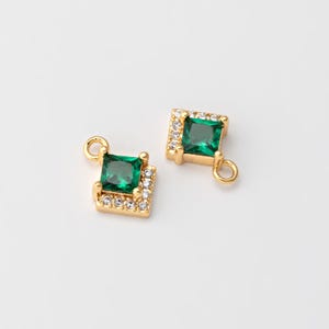 May include: Two gold-colored square charms with a central emerald-green square gemstone. Each charm is surrounded by small, clear gemstones and has a small loop for attachment. The charms are set against a plain white background.