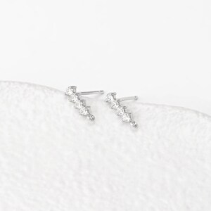 Puede incluir: Un par de pendientes de botón plateados. Cada pendiente presenta una serie de cinco gemas redondas y transparentes dispuestas en línea curva. Se encuentran sobre un fondo blanco.