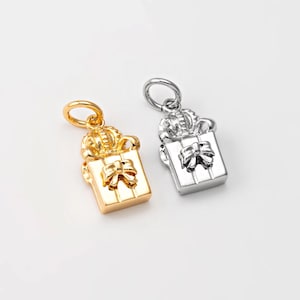 May include: Two gift box charms, one gold and one silver, are shown against a white backdrop. Each charm has a bow and a decorative top. Ideal for jewellery making or crafting projects.