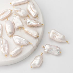 Op de afbeelding: Onregelmatig gevormde, witte zoetwaterparels met een glanzende glans. Elke parel is afzonderlijk bedraad met een goudkleurige lus voor het maken van sieraden. De parels variëren in grootte en vorm, sommige zijn langwerpig, andere ronder.