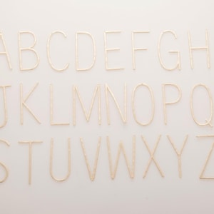 Puede incluir: Alfabeto de la A a la Z en letras doradas, dispuestas sobre un fondo blanco. Las letras están en una fuente sencilla y parecen estar hechas de un material texturizado. Las letras están espaciadas uniformemente, creando una exhibición decorativa.