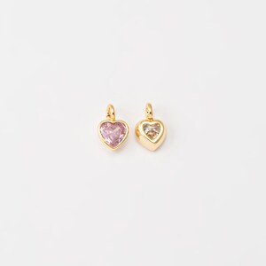 May include: Two gold heart-shaped charms with clear crystal accents. The charm on the left has a pink crystal, while the charm on the right has a clear crystal.