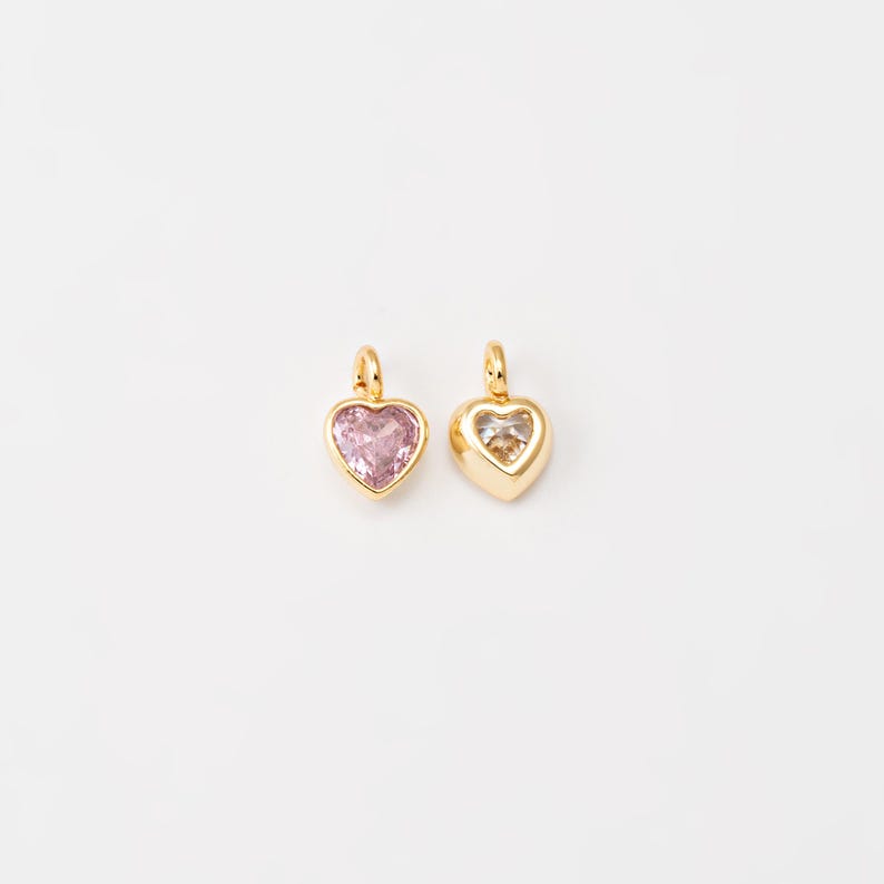 May include: Two gold heart-shaped charms with clear crystal accents. The charm on the left has a pink crystal, while the charm on the right has a clear crystal.
