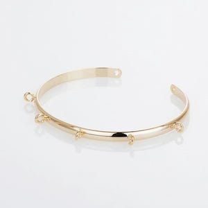May include: A gold-colored open cuff bracelet with multiple small loops for attaching charms. The bracelet has a smooth, polished finish and a simple, elegant design. The bracelet is shown on a white background.