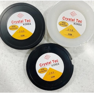 Może przedstawiać: Trzy szpule sznurka do koralików marki Crystal Tec. Szpule są czarne i białe, z żółtymi i białymi etykietami. Etykiety wskazują średnicę sznurka (0,3, 0,5 i 1,0) oraz długość (50 m). Widoczny jest również tekst "NEW STRONG".