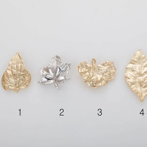 May include: Four metal leaf charms in gold and silver tones. The charms are in various leaf shapes, including a teardrop, maple leaf, and two other textured leaf designs. The charms are arranged on a white background.