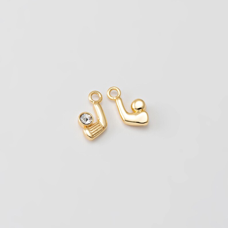 May include: Two gold-colored golf-themed charms. One charm features a golf club with a clear crystal accent, and the other depicts a golf ball. Both charms have a small loop at the top for attaching to jewelry.