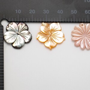 Puede incluir: Tres colgantes con forma de flor. Los colgantes son de n&aacute;car. El primero es de color gris oscuro, el segundo dorado y el tercero rosa claro. Cada colgante tiene un agujero en la parte superior para ensartar. Medidas en cent&iacute;metros.
