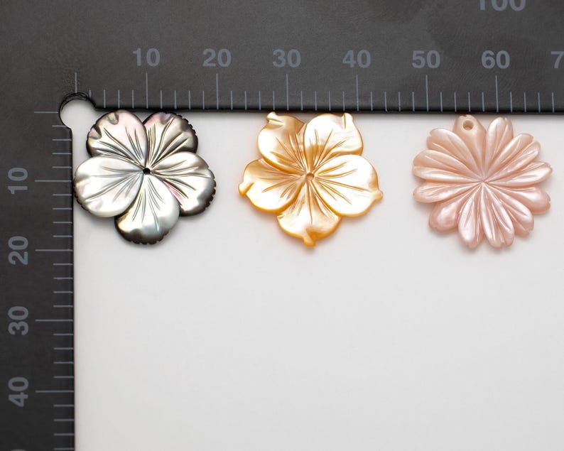 Puede incluir: Tres colgantes con forma de flor. Los colgantes son de n&aacute;car. El primero es de color gris oscuro, el segundo dorado y el tercero rosa claro. Cada colgante tiene un agujero en la parte superior para ensartar. Medidas en cent&iacute;metros.