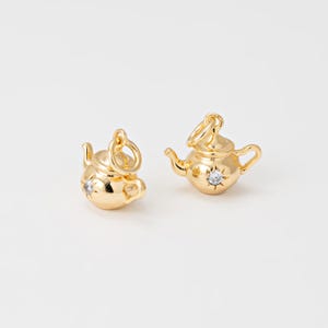 May include: Two gold-colored teapot charms with a star-shaped crystal accent. The charms have a loop at the top for attaching to a necklace or bracelet. The teapots have a handle, spout, and lid.