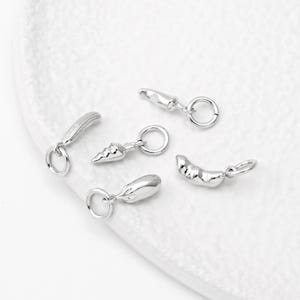 Könnte beinhalten: Fünf silberne Anhänger in Form von Gemüse, darunter eine Karotte, eine Aubergine, eine Gurke, eine Paprika und eine Bohnenschote. Jeder Anhänger hat einen Sprungring zum Anbringen an einem Armband oder einer Halskette.