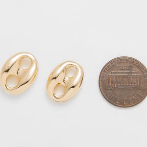 May include: Two gold-colored, bean-shaped charms with two holes each, next to a US one-cent coin. The charms have a shiny, reflective surface. The coin shows the US Capitol building and the words "United States of America."