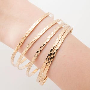 Può includere: Una collezione di braccialetti color oro con una texture martellata. I braccialetti sono impilati su un polso, mostrando il loro design semplice ma elegante. La luce si riflette sulla superficie strutturata.