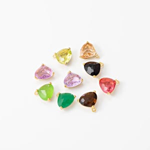 Puede incluir: Una colección de colgantes triangulares de piedras preciosas de colores con detalles metálicos dorados. Las piedras preciosas son de varios tonos, incluyendo amarillo, rosa, verde y marrón. Cada colgante tiene un lazo para sujetar.