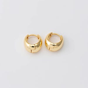 May include: Two gold-coloured, chunky hoop earrings. Each earring has a rounded, polished design with a hinged closure. The earrings are set against a plain white background, highlighting their shiny, metallic appearance. These earrings are a classic jewellery accessory.