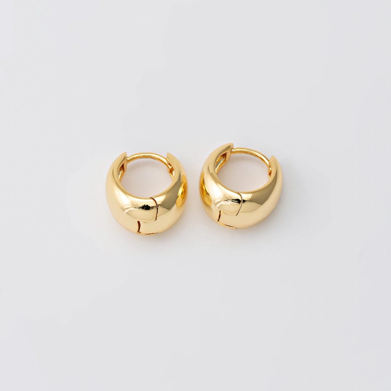 May include: Two gold-coloured, chunky hoop earrings. Each earring has a rounded, polished design with a hinged closure. The earrings are set against a plain white background, highlighting their shiny, metallic appearance. These earrings are a classic jewellery accessory.